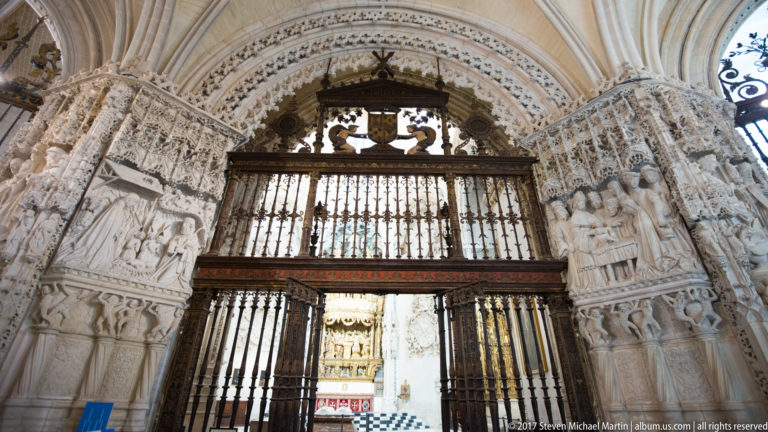 Ornately carved stone chapel entrance with a tall dark iron grille and heraldic crest above.