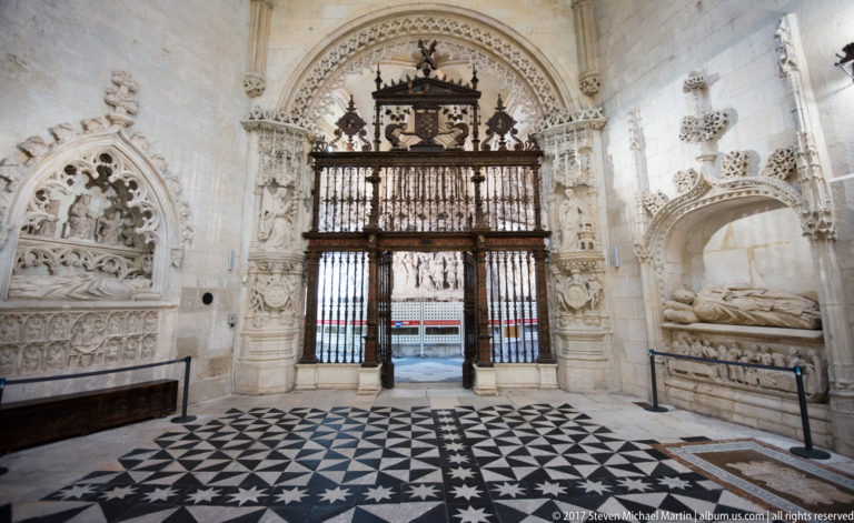 Pale-stone cathedral chapel with iron screen, wall tomb, carved portal, and black-and-white patterned floor.
