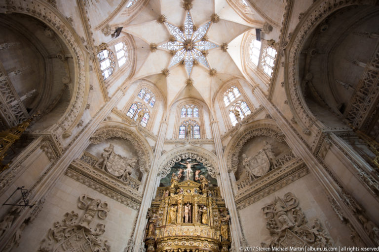 Upward view of an elaborate star-ribbed Gothic vault above windows, stone monuments, and a gilded altarpiece.