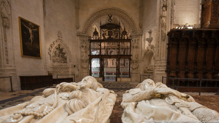 Paired pale-stone tomb effigies viewed toward a dark iron chapel screen and carved choir-like woodwork.
