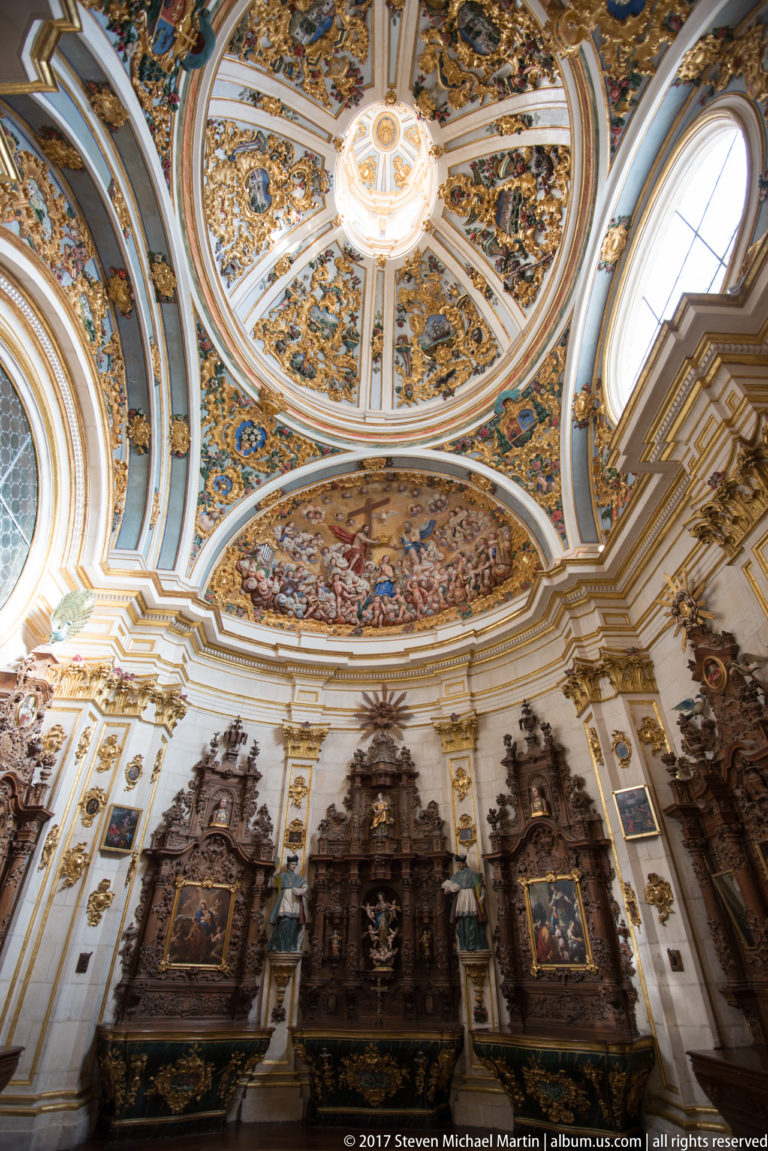 Baroque chapel with dark carved retables, framed paintings, oval windows, and a richly painted gilded dome.