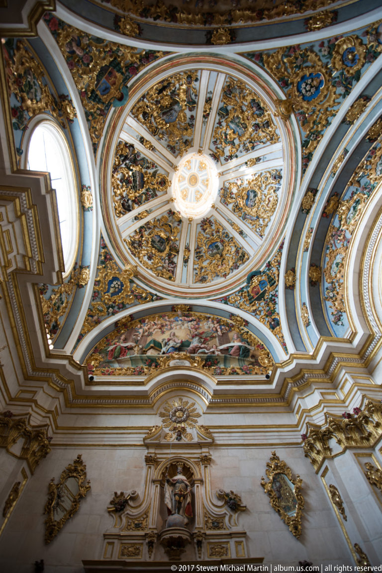 Symmetrical upward view of a segmented painted dome with gilded ornament, central medallion, and oval windows.