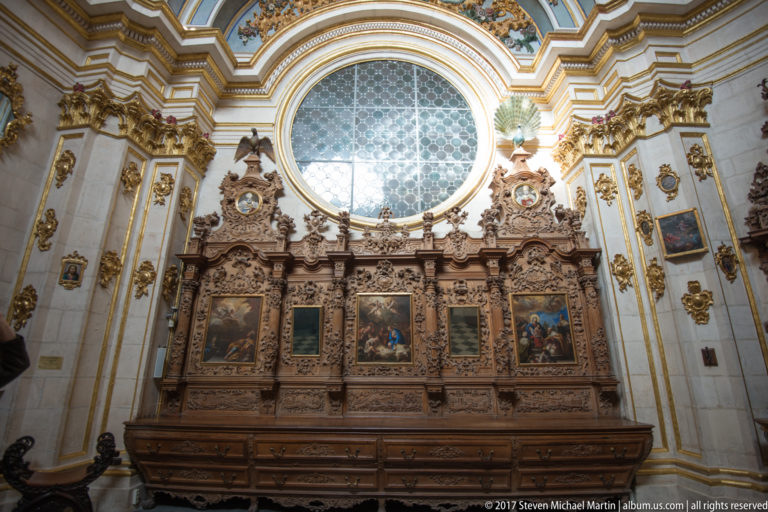 Long carved dark-wood furnishing with small religious paintings beneath a large oval window in a gilded chapel.