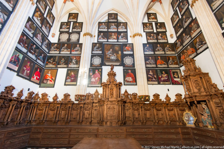 Oblique view of carved choir stalls beneath tiered portraits of clergy extending around a vaulted room.