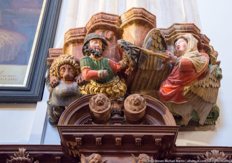 Polychrome carved bearded man in a broad hat flanked by two winged figures atop dark woodwork.