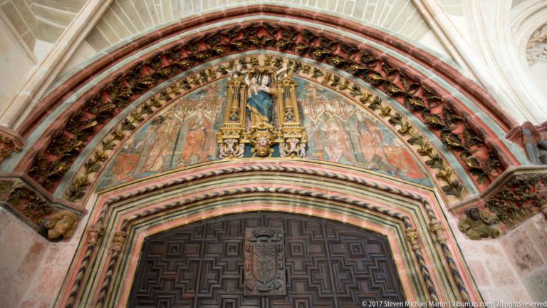 Pointed medieval doorway with painted carved archivolts, polychrome tympanum, and dark paneled doors.