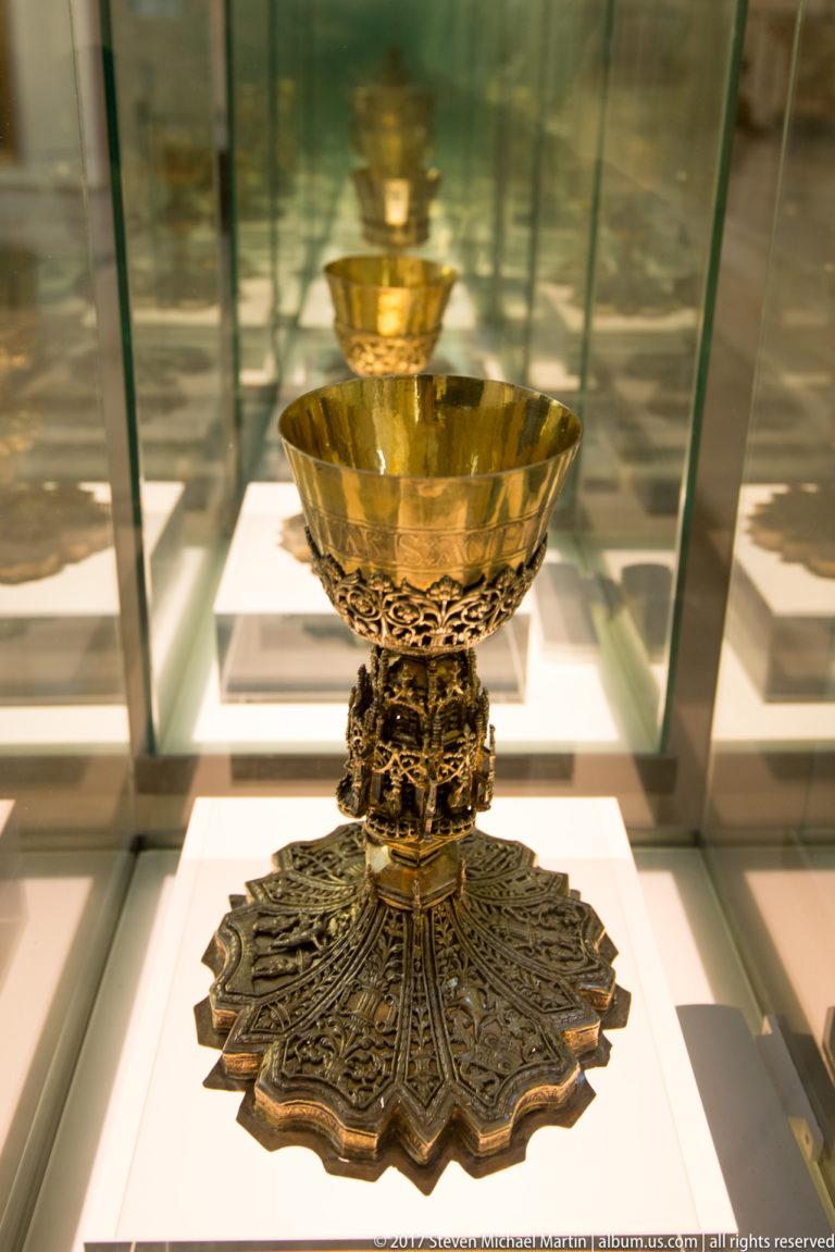 Ornate metal liturgical vessel with shallow bowl, pierced stem, and broad lobed decorated foot in a glass case.