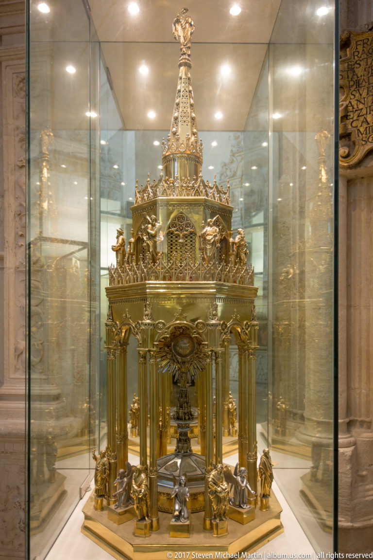 Gilded miniature Gothic-style shrine with columns, arches, figures, pinnacles, and tall spire inside a glass case.