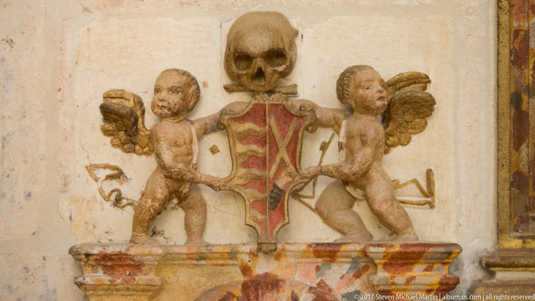 Skull above a red heraldic shield held by two winged putti on a pale carved wall relief.