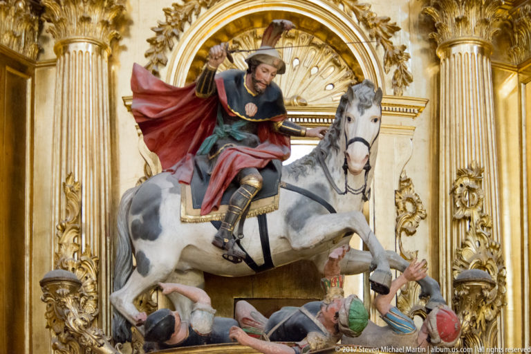 Polychrome saint on a white horse above fallen figures in a gilded niche with red cloak and raised arm.