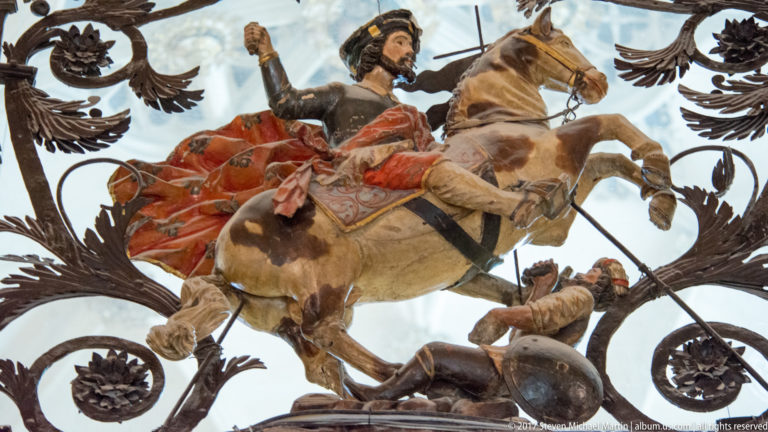 Polychrome mounted warrior in a red cloak riding through dark scrolling ironwork with smaller figures below.