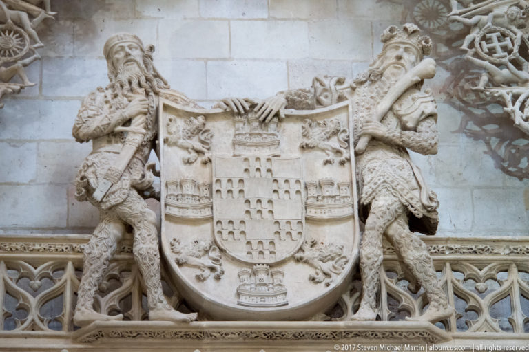 Large pale-stone coat of arms supported by two carved armored figures above a Gothic tracery band.