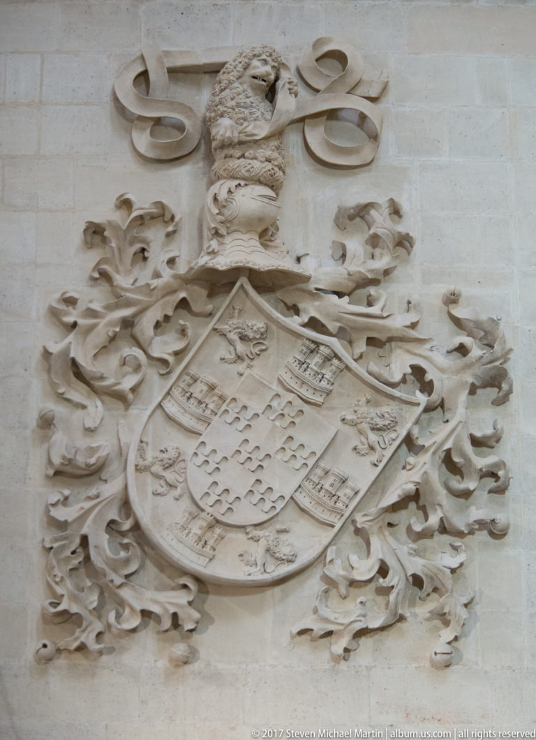 Pale-stone quartered coat of arms beneath a crested helmet, surrounded by curling carved mantling.