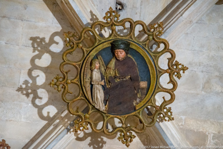 Gilded circular medallion with painted religious figures mounted at the intersection of pale stone vault ribs.