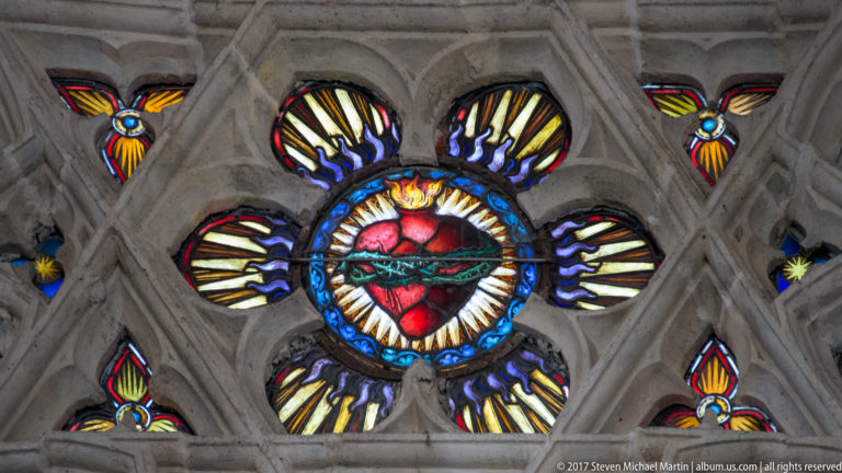 Red Sacred Heart stained glass centered in a small Gothic tracery window with blue, yellow, and red surrounding panes.