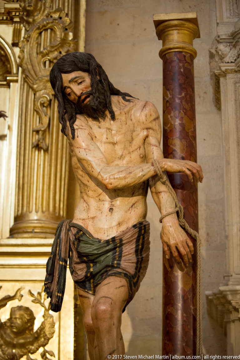 Life-sized polychrome Christ with lowered head, bound hands, wounds, and a column beside him.