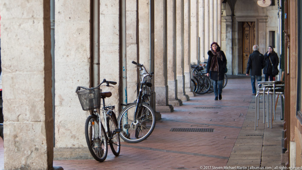 Bicycles parked beneath a long stone arcade as pedestrians walk past cafés and shopfronts in Burgos.