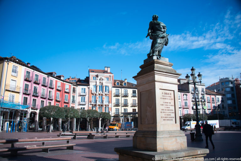 Bronze statue on a tall pedestal in Plaza Mayor, surrounded by colorful façades, trees, and streetlamps.