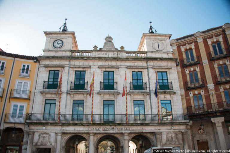 Symmetrical Burgos City Hall façade with central clock, roof balustrade, flags, balconies, and ground-floor arches.