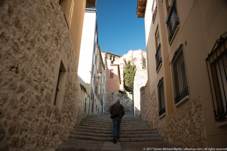 Narrow stone stairway climbing between old masonry houses, barred windows, and a lone pedestrian in Burgos.