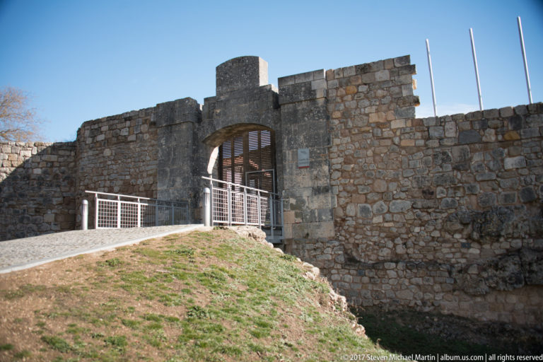 Ruined stone walls and reconstructed arched gateway at Burgos Castle, with railings and a grassy slope in front.