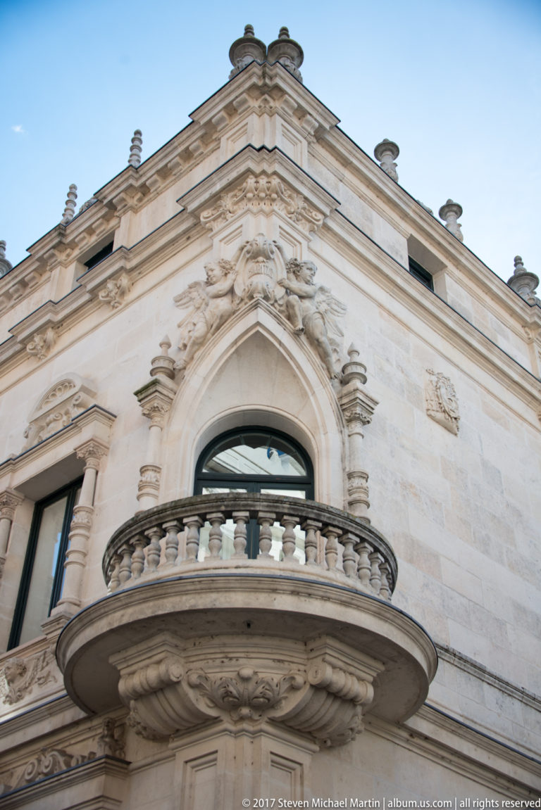 Upward view of a semicircular stone balcony beneath an arched window on an ornate pale-stone façade.