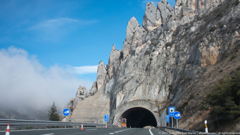 Highway entering a tunnel beneath jagged gray limestone cliffs, with road signs and barriers in the foreground.