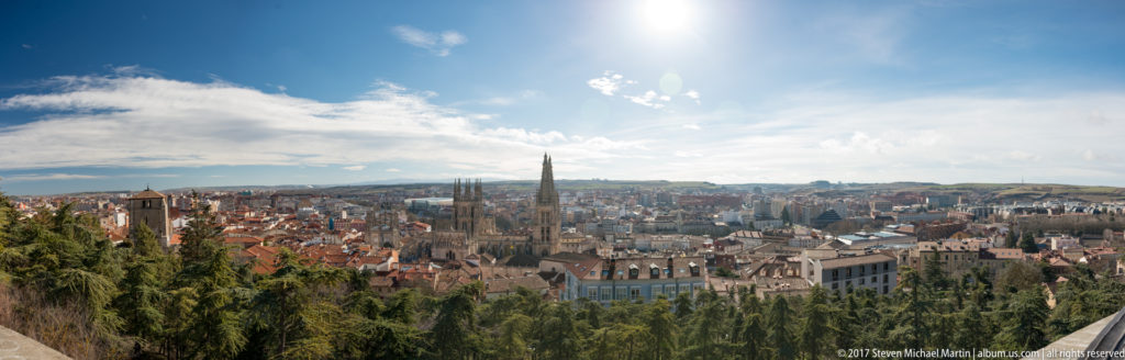 Wide elevated panorama of Burgos with cathedral spires near the center, foreground trees, dense rooftops, and distant hills.