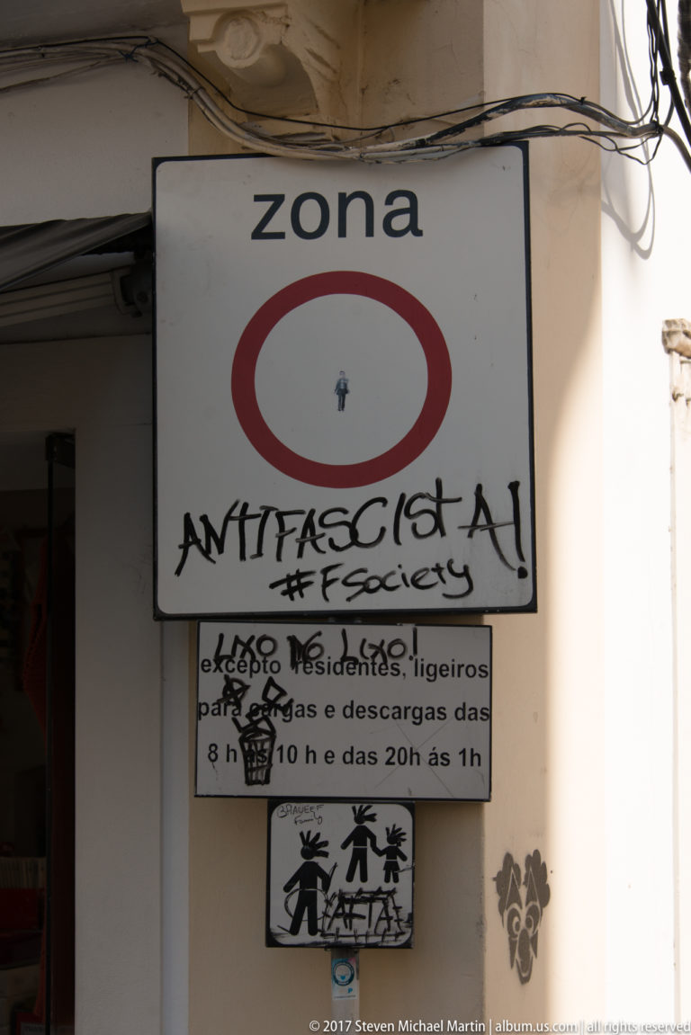 Graffiti-covered “zona” traffic sign and smaller notices mounted beside a shaded doorway in Coimbra.