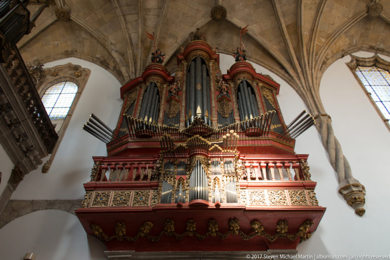 Large red-and-gold pipe organ with silver pipes and horizontal trumpets beneath a ribbed stone vault.