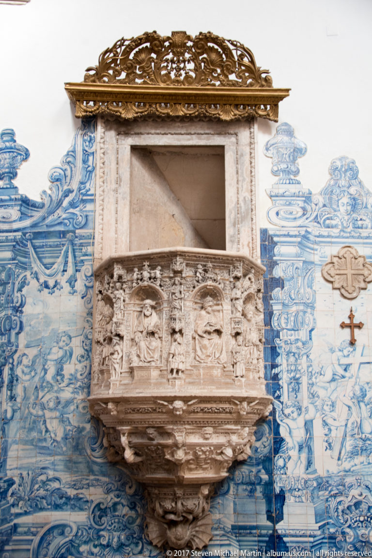 Carved pale-stone pulpit with relief figures and canopy against blue-and-white azulejo tiles.