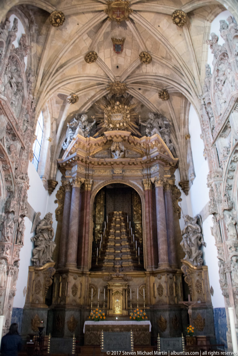Monumental carved tomb structure behind an altar, framed by dark columns beneath a ribbed vault.