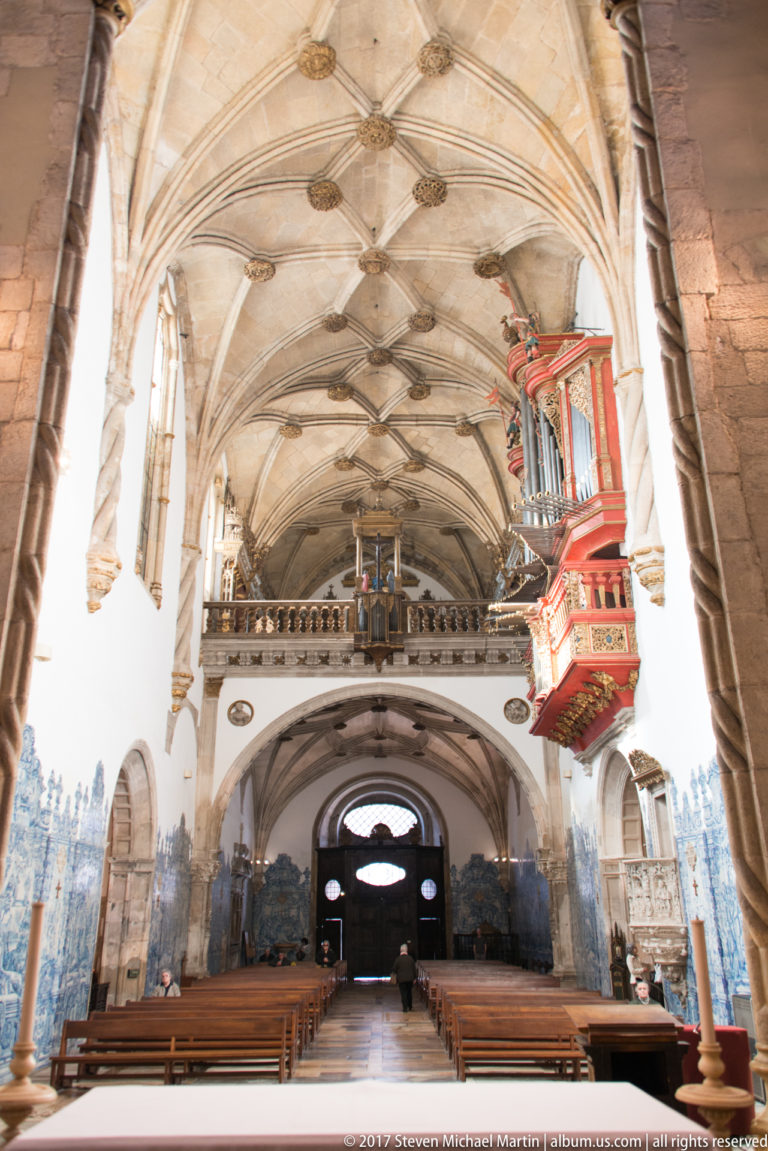 Rear-facing view down Santa Cruz Church nave with pews, tiled walls, ribbed vaults, and red organ.