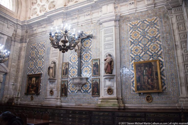 Ornate wall with blue-and-gold tiles, framed religious paintings, saint statues, chandeliers, and dark cabinetry.
