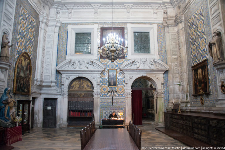 Symmetrical tiled wall with two carved doorways, tall windows, chandelier, paintings, and dark cabinetry.