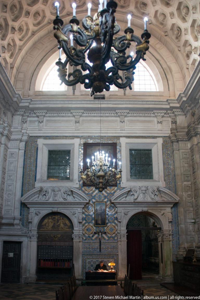 Two dark chandeliers hanging before a high arched window, carved stonework, and blue-and-gold tiled walls.
