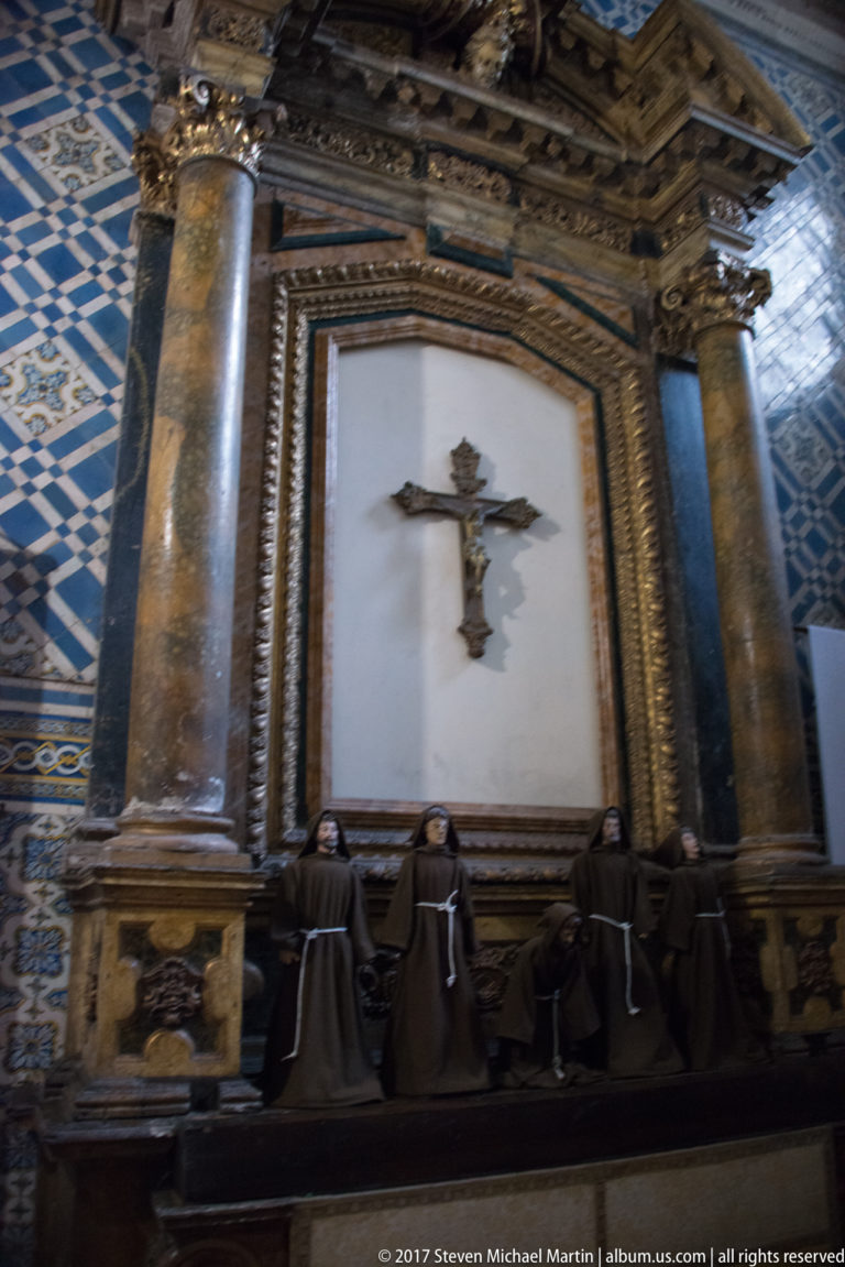 Crucifix in a dark columned frame above kneeling robed figures, with blue patterned tiles at the sides.