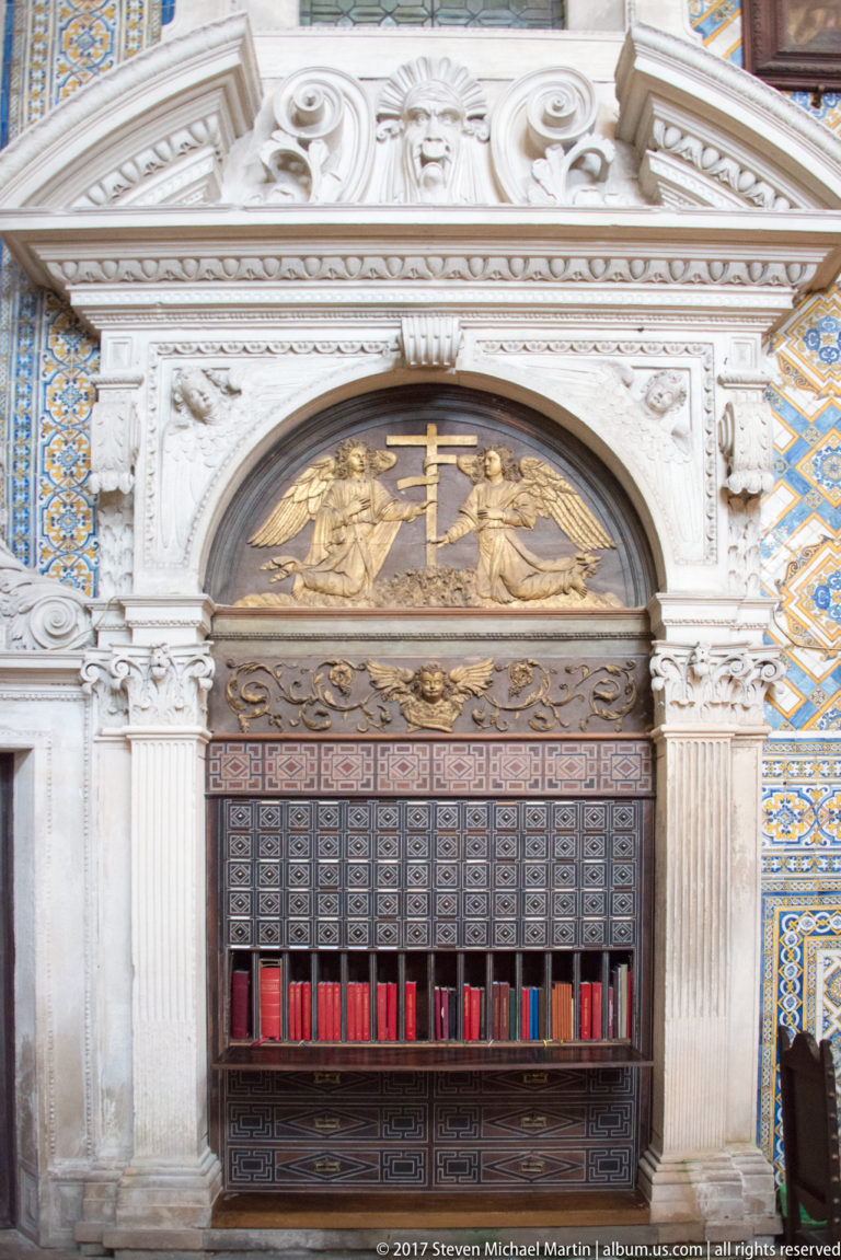 Carved white-stone surround with cross relief above a dark cabinet of small compartments and red volumes.