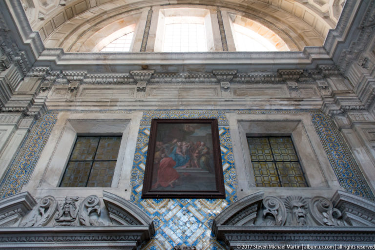 Upward view of tall carved stone wall with blue-and-gold tiles, windows, and a central religious painting.