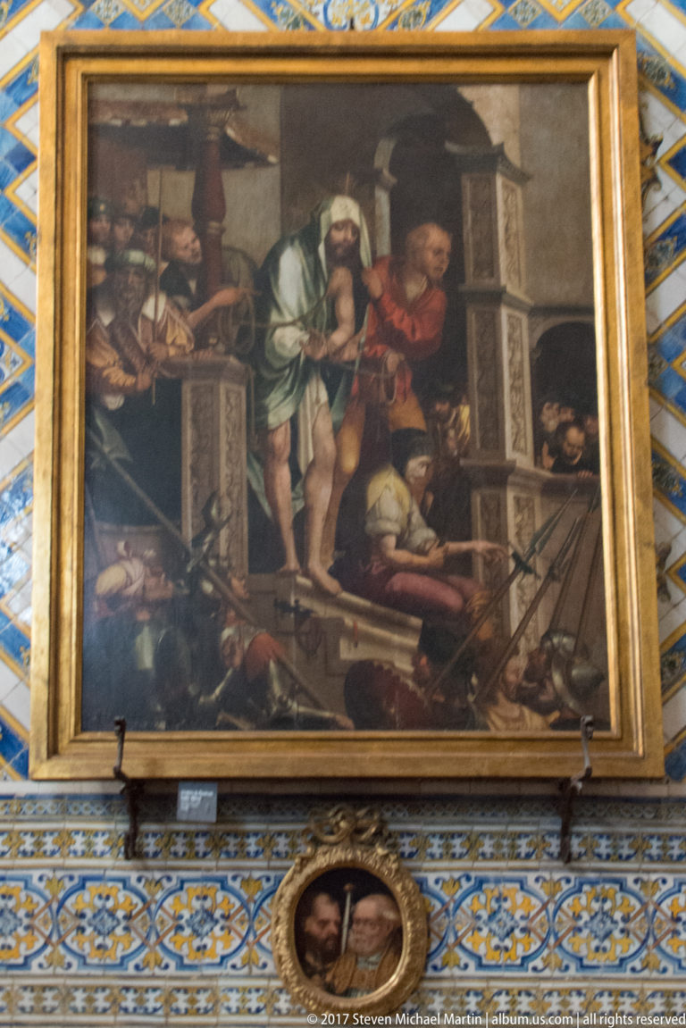 Gilded rectangular religious painting with many figures around an architectural scene, mounted on patterned tiles.