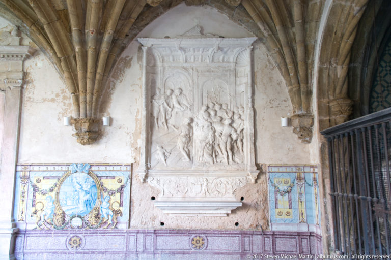 Weathered stone relief above colorful tiles beneath ribbed cloister vaulting, with an iron gate at right.