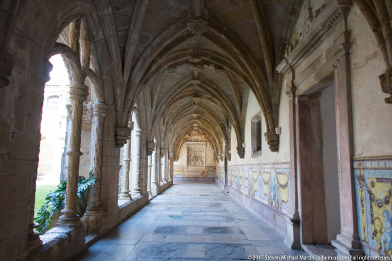 Long rib-vaulted cloister corridor with open arcade at left and colorful tiled wall panels at right.