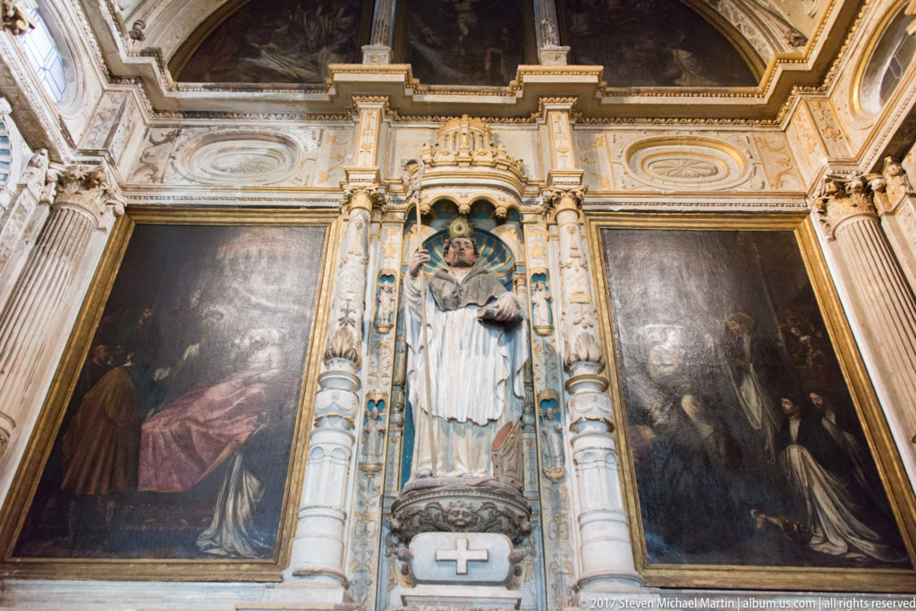 Ornate altarpiece with central saint statue, flanking religious paintings, columns, gilding, and painted decoration.