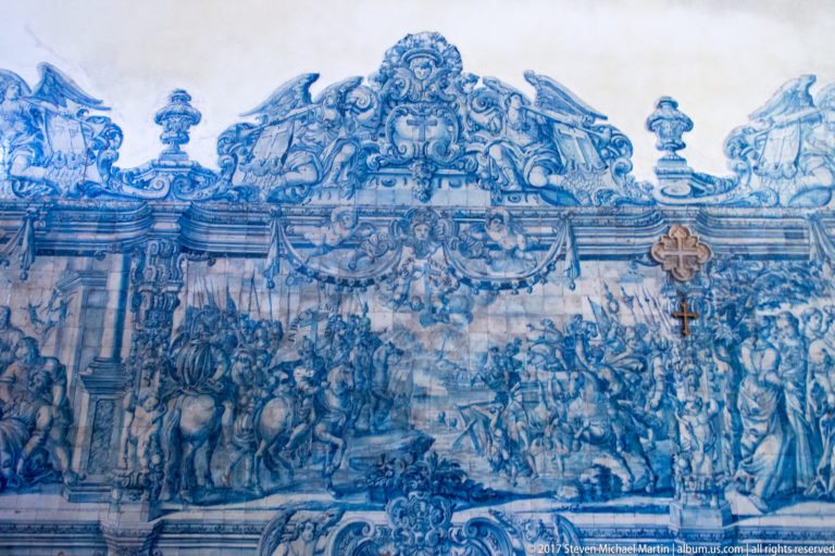 Large blue-and-white azulejo panel with crowded narrative scene and elaborate scrolling ornamental border.