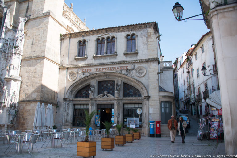 Historic arched café-restaurant façade with outdoor tables and pedestrians on a square in central Coimbra.
