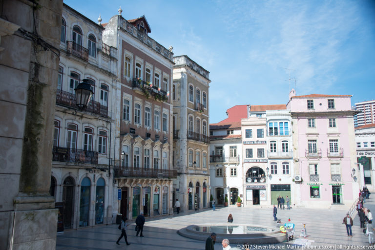 Pedestrians cross a paved Coimbra square bordered by tall historic façades, balconies, pastel buildings, and a low circular fountain.