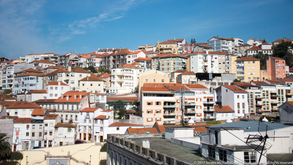 Dense white and pastel buildings with red tile roofs rise across a steep hillside under a clear blue sky in Coimbra.