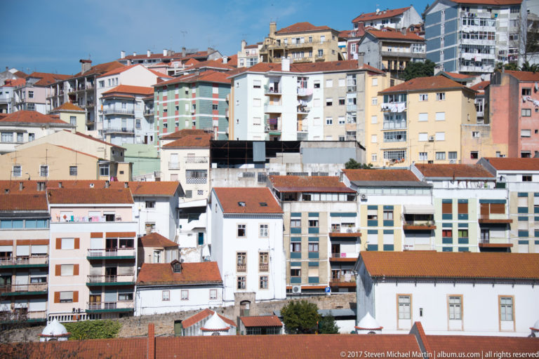 Closely packed apartment blocks and older tiled-roof houses step up a steep Coimbra hillside in white, ocher, green, and pink.