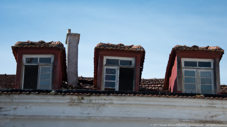 Three weathered dormer windows and a masonry chimney rise from an old red-tile roof against blue sky.