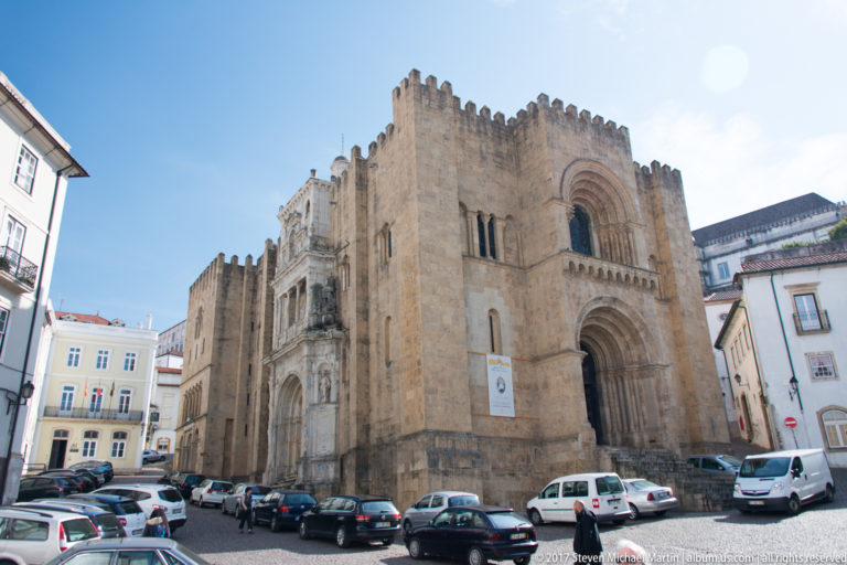Fortress-like stone west façade of Coimbra’s Old Cathedral with crenellations, recessed Romanesque portal, and cars along the sloping street.